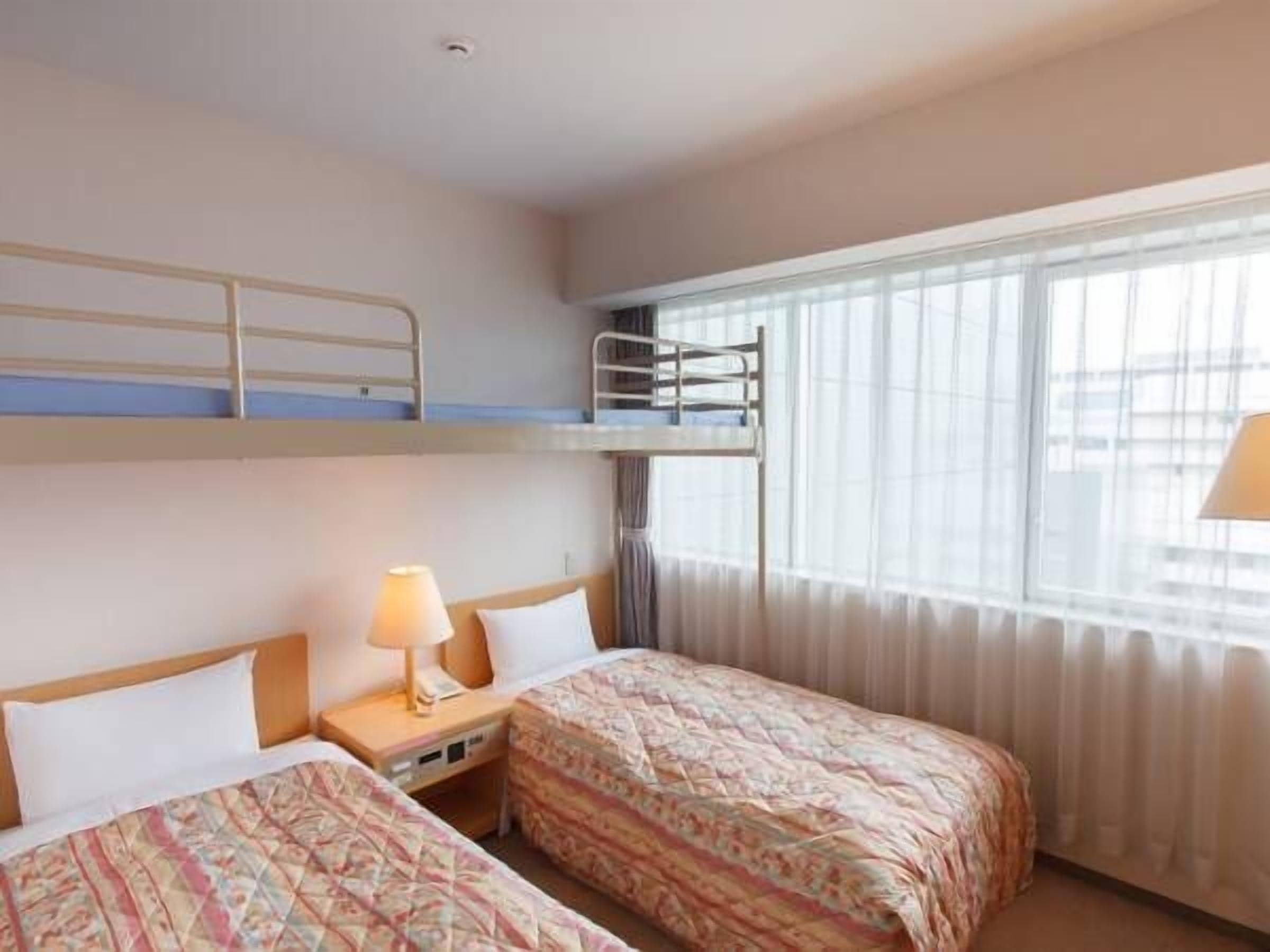 Hotellihuone, jossa on kaksi sänkyä, kerrossänky, yöpöytä lampun kanssa ja ikkuna, jossa on verhot.