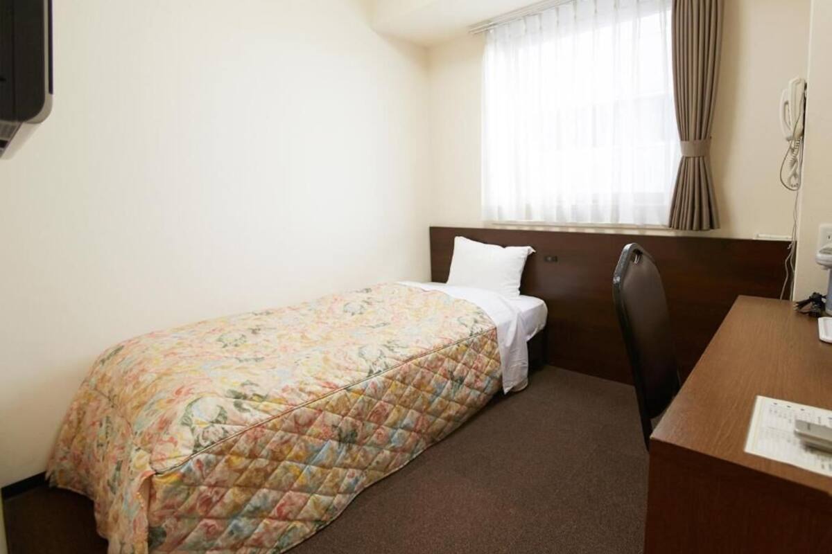 A small hotel room with a single bed, a wooden headboard, a desk with a chair, and a window with curtains.