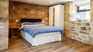 A bedroom with a stone wall, a bed with a blue and white bedspread, a wooden floor, and a window with a view of the outside. - CROSS HOUSE STUDIO Dog Friendly Stone Barn Conversion in the Heart of Warkworth (Warkworth)
