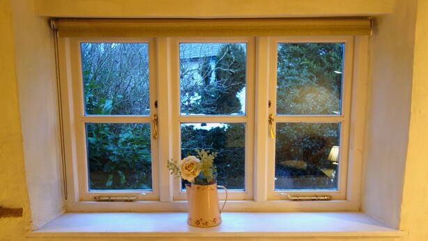 A window with three panes, a vase with flowers on the ledge, and a view of trees and a building outside. - Charming, secluded, characterful cottage in the heart of Lechlade (Lechlade on Thames)