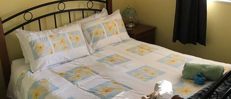 Una cama con ropa de cama floral, una mesita de noche con lámpara, un cartel en la pared y una ventana con cortinas.
