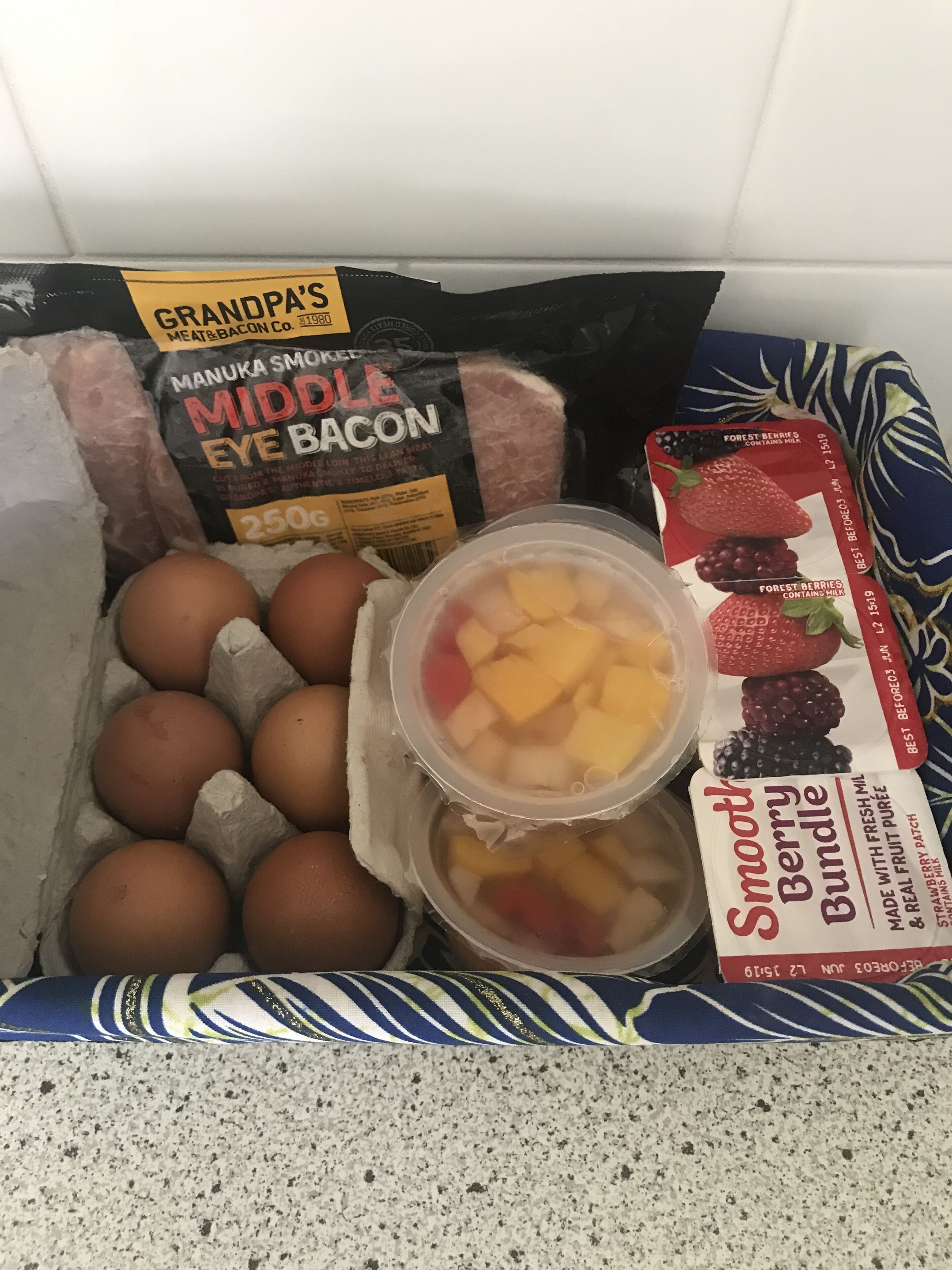 Una caja con huevos, un paquete de bacon de ojo central ahumado con miel de Manuka y un recipiente con frutos rojos cremosos.