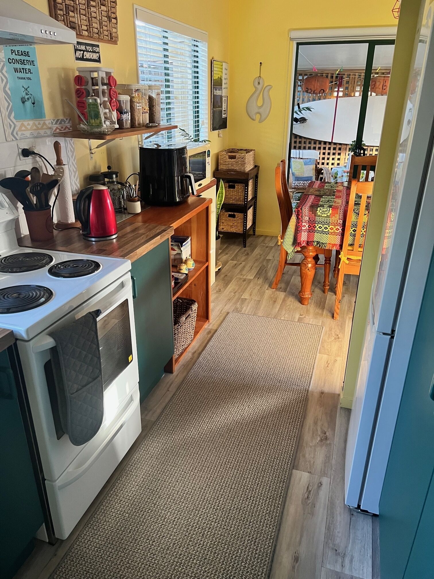 Et køkken med komfur, elkedel og kaffemaskine. Der er et spisebord med stole og en måtte på gulvet.