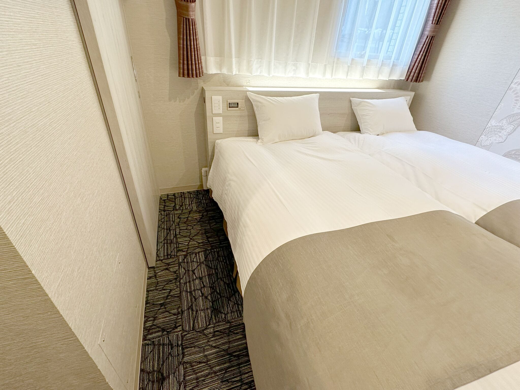Una habitación de hotel con cama, mesita de noche, toma de corriente y ventana con cortinas de gasa.