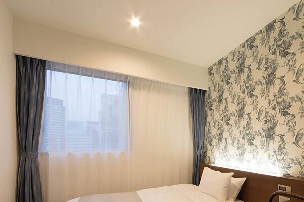 A hotel room with a bed, a chair, a window with curtains, and a floral wallpaper.