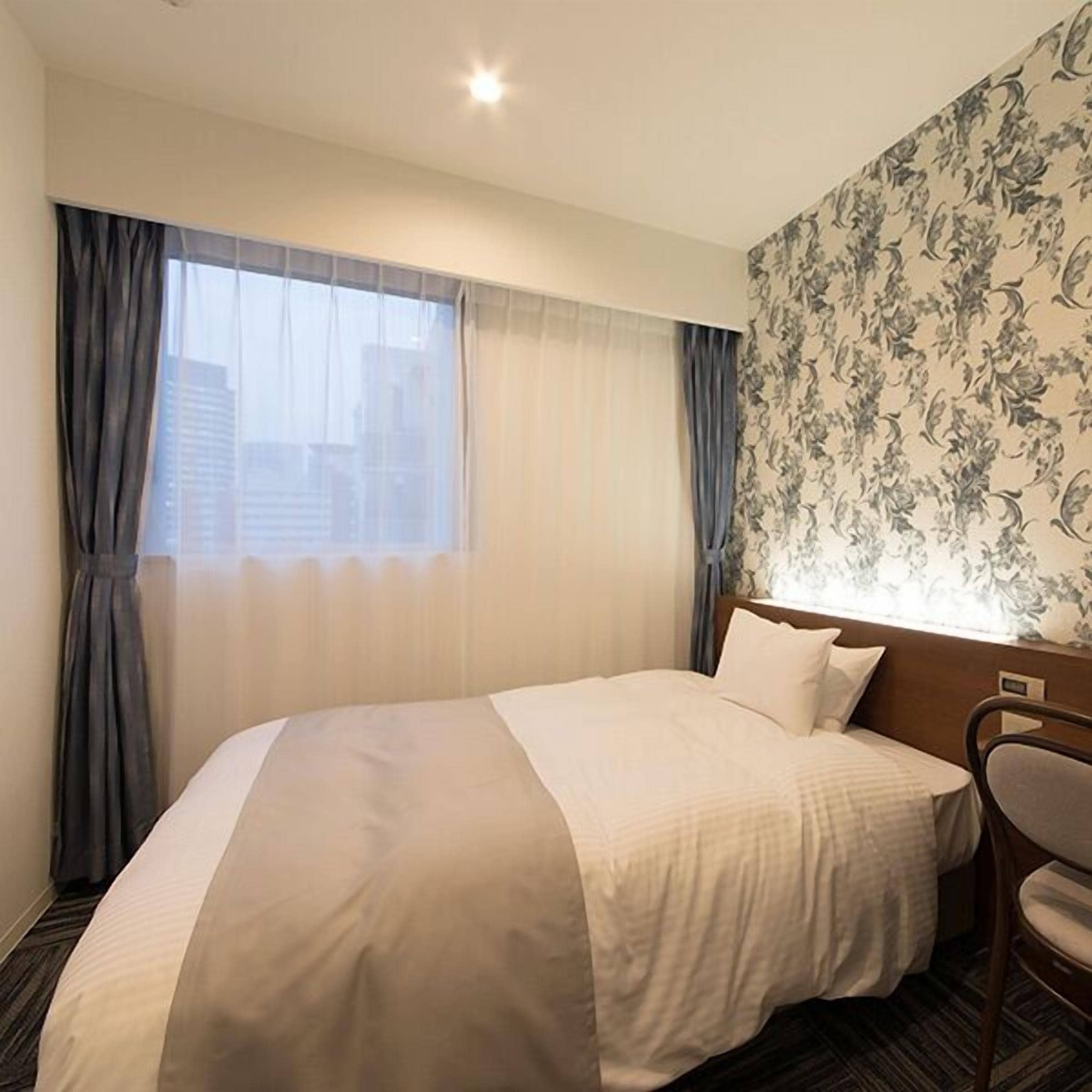 A hotel room with a bed, a chair, a window with curtains, and a floral wallpaper pattern.