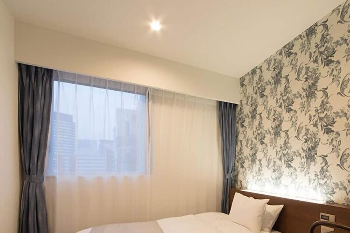 Una habitación de hotel con cama, una silla, un ventanal con cortinas y un estampado floral en el papel tapiz.