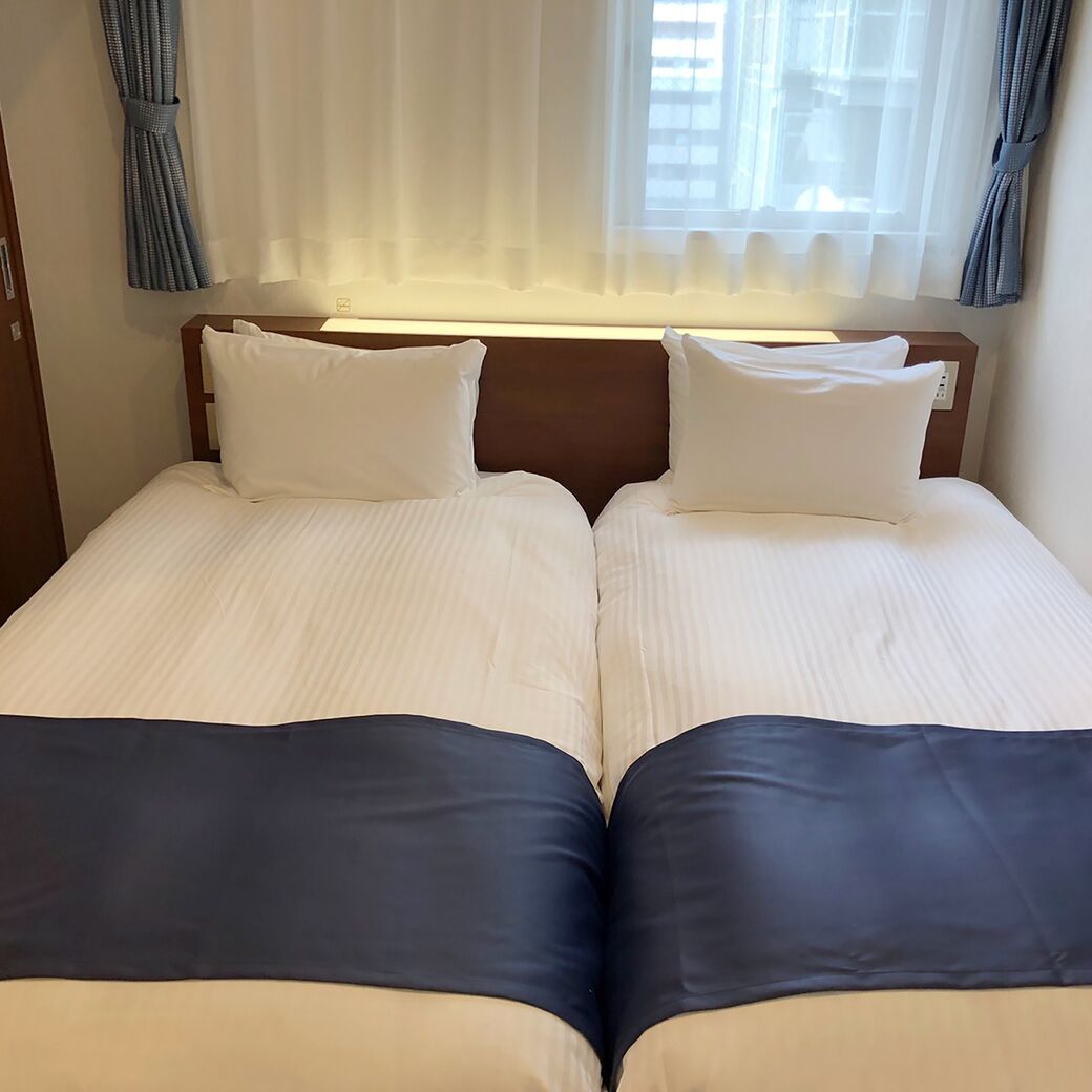 A double bed with white linens and dark blue bedspreads, a wooden headboard, and sheer curtains.