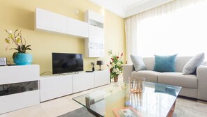 Een moderne woonkamer met een flatscreen televisie, een glazen salontafel, een bank met kussens en een potplant.