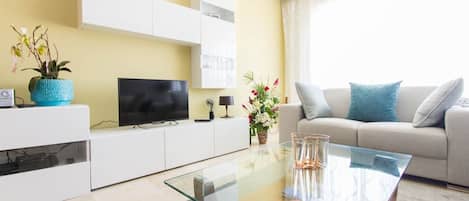 Una sala de estar moderna con una televisión de pantalla plana, una mesa de centro de vidrio, un sofá con cojines y una planta en maceta.