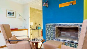Een woonkamer met twee beige fauteuils, een klein houten tafeltje met een schaakbord, een blauwe betegelde open haard en een keuken op de achtergrond.