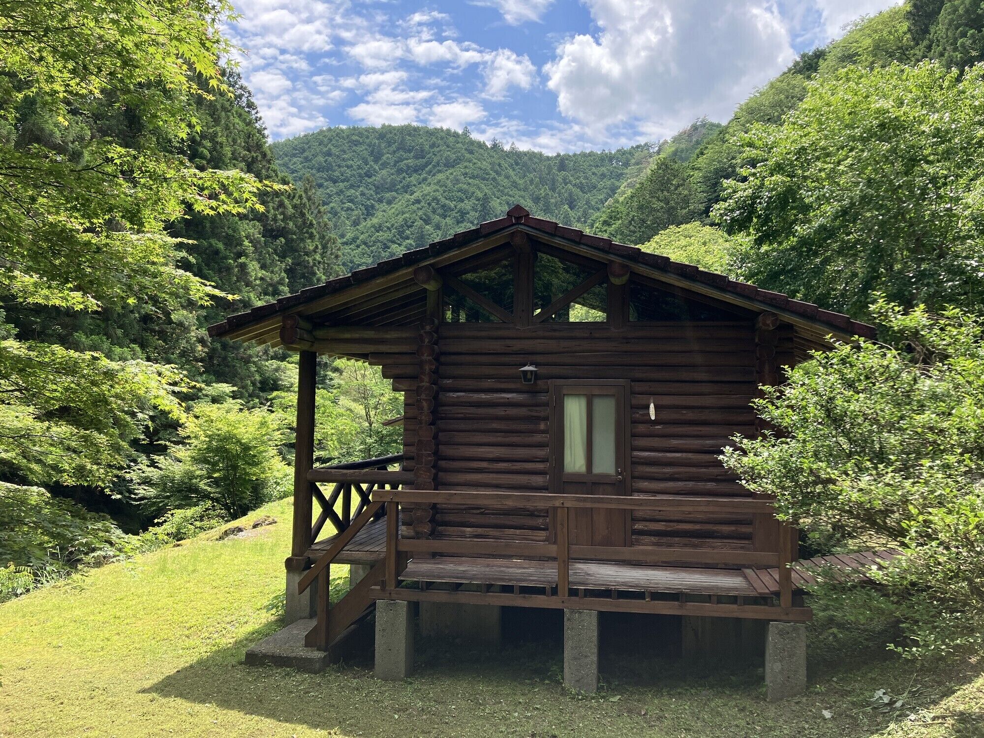 A wooden cabin with a porch, surrounded by lush greenery and a mountain backdrop.