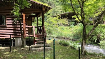 A wooden cabin with a porch, surrounded by lush greenery and a small stream.