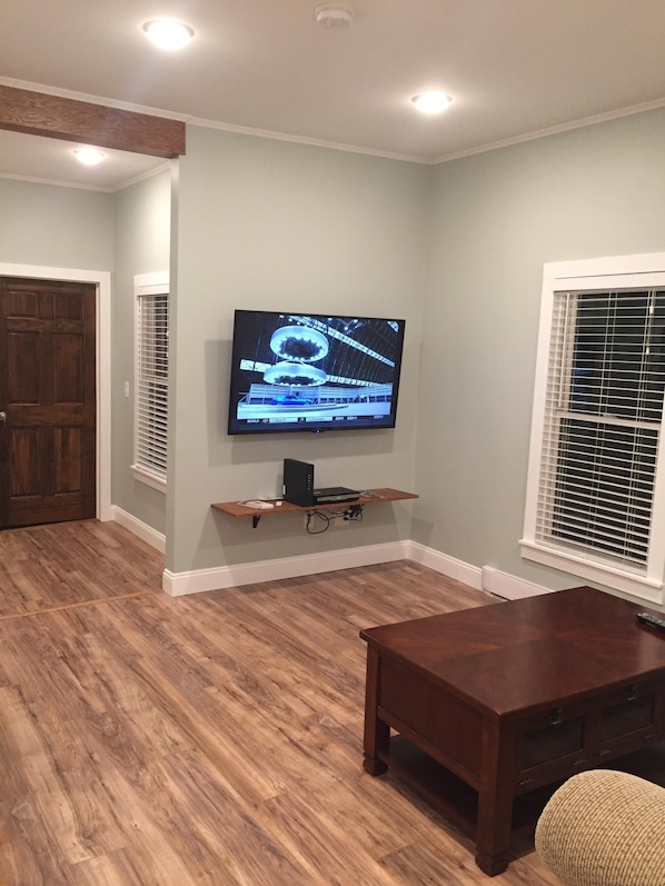 Una sala de estar con un televisor de pantalla plana montado en la pared, una mesa de centro de madera y una ventana con persianas.