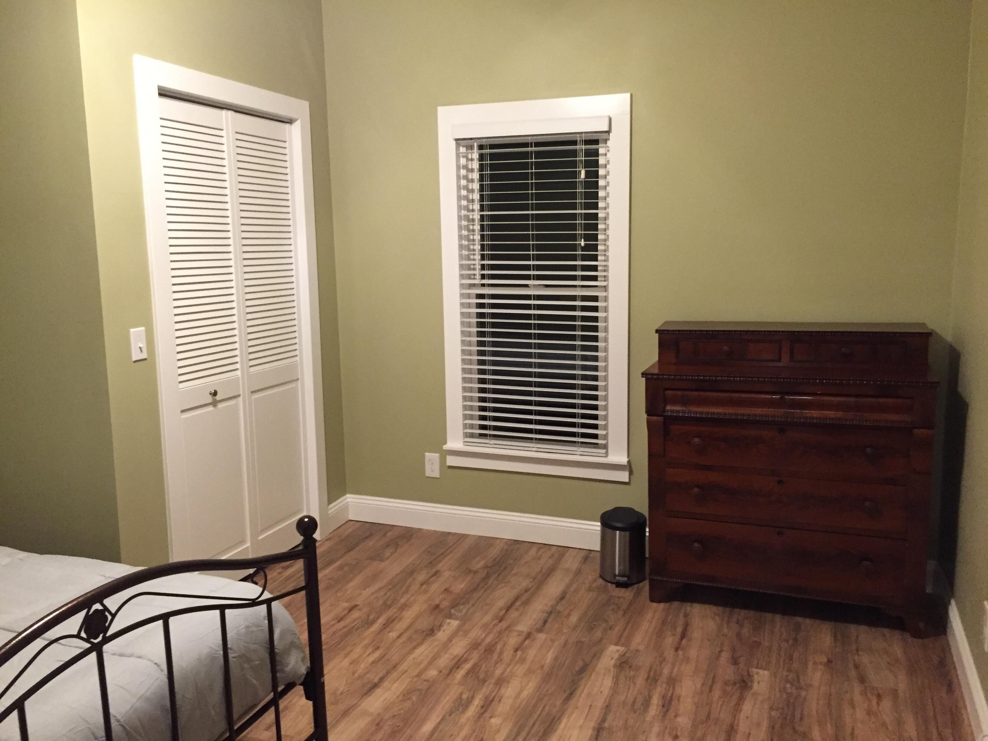 A room with a bed, a dresser, a window with blinds, and a trash can.