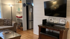 Una sala de estar con suelo de madera, un sofá, una mesa de centro, un televisor sobre un mueble bajo, y una cocina al fondo.