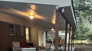 Un patio al aire libre con suelo de ladrillo, una zona cubierta con sillas y una mesa, y un muro de ladrillo.