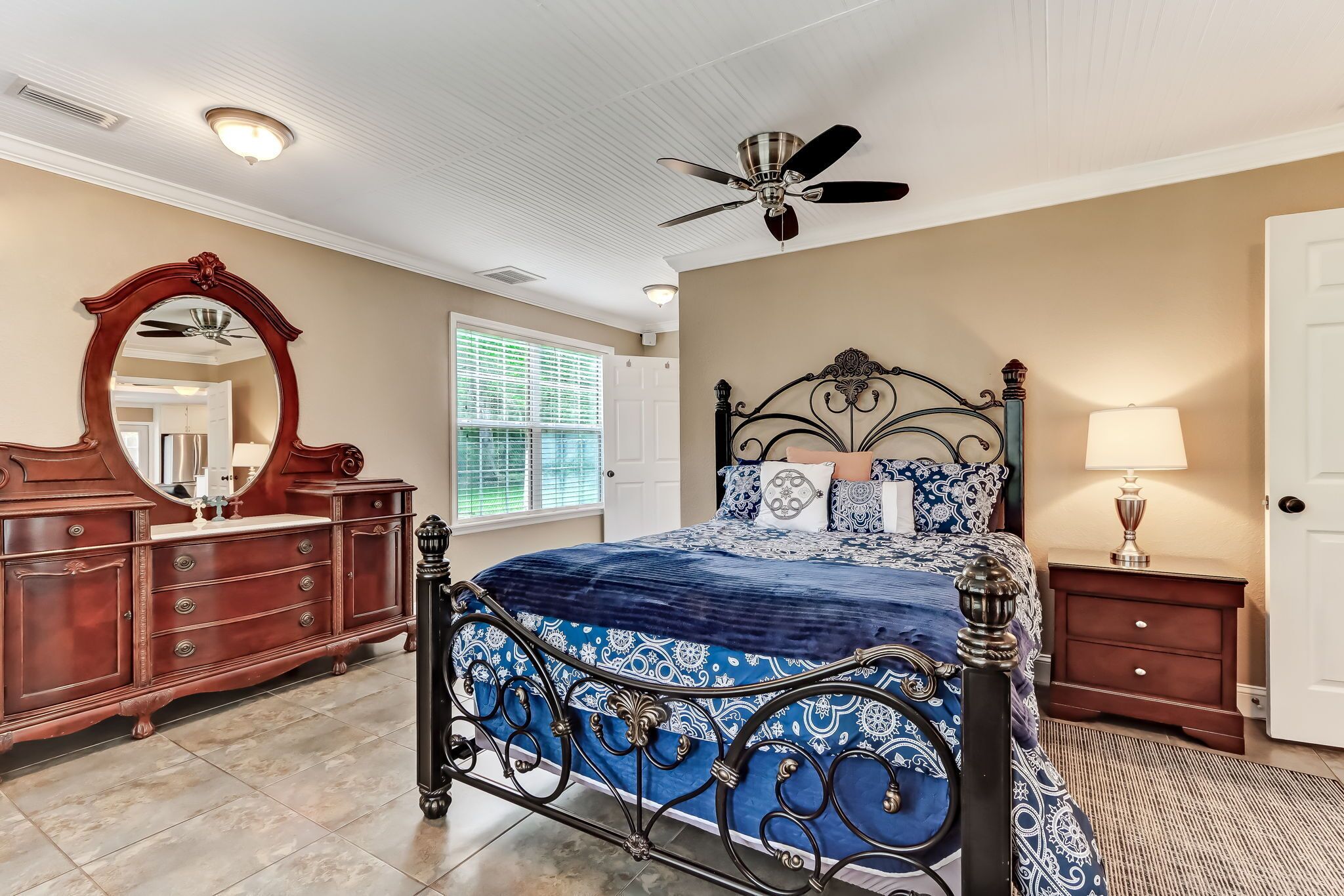 Une chambre à coucher avec un grand lit, une commode, un miroir, un ventilateur de plafond et une fenêtre avec des stores.