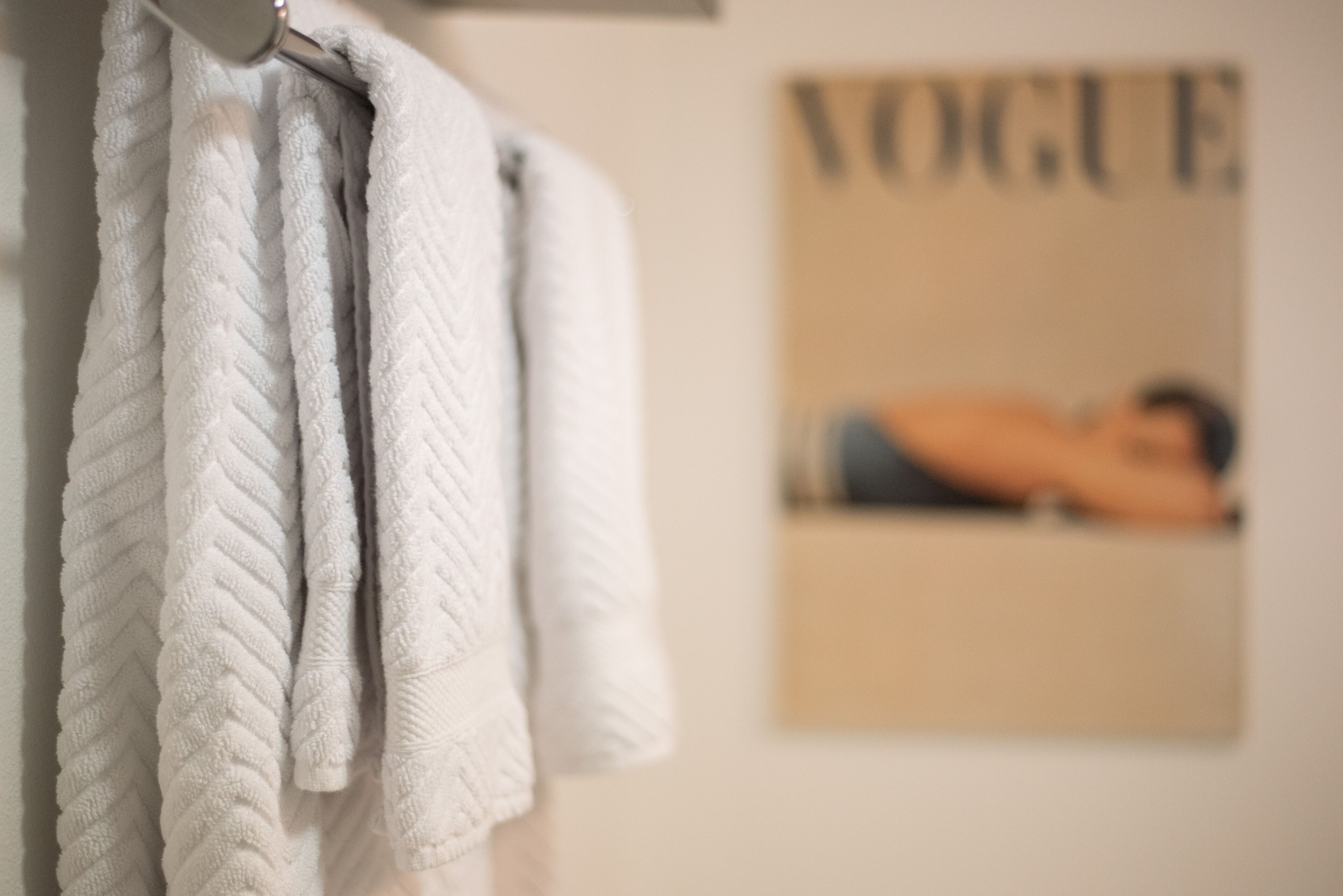 A bathroom with a towel rack holding several towels. In the background, there is a poster on the wall with the word "VOGUE" visible.