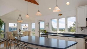 Una cocina moderna con una isla grande, iluminación colgante y amplios ventanales que ofrecen vista al mar.