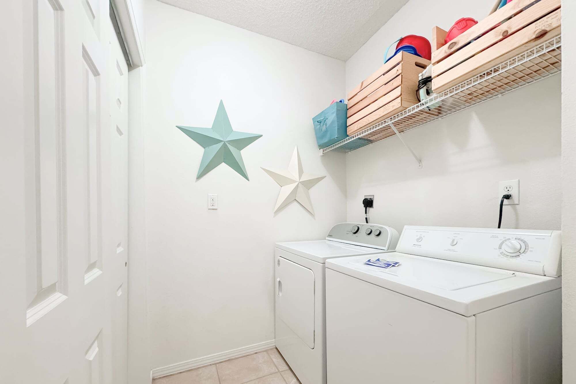 A laundry room with two white front-loading washing machines, a wooden shelf above holding various items, and two star-shaped wall decorations.