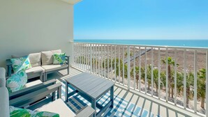 Varanda com sofá e mesa, vista para uma praia com palmeiras e céu claro.