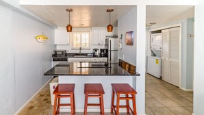 Una cocina moderna con una isla central, taburetes de bar, iluminación colgante y vistas al área de lavandería contigua.