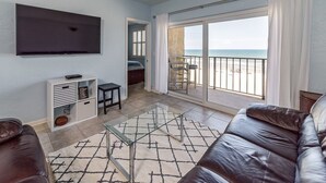 Una sala de estar con un televisor grande, una mesa de centro de vidrio, un sofá de cuero marrón y vistas a la playa a través de un cristalero corredizo.