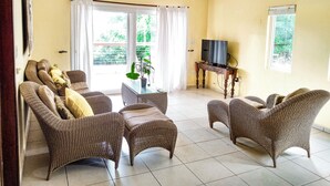 Ein Wohnzimmer mit Rattanmöbeln, Deckenventilator, Fernseher auf einem Holzboden und Blick ins Grüne.