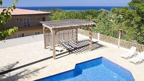 Ein Swimmingpool mit einer Pergola und Liegestühlen, umgeben von einer gepflasterten Terrasse und Blick auf den Ozean.