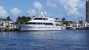 Eine große weiße Yacht liegt in einer Marina vor Anker, im Hintergrund sind weitere Boote und ein hohes Gebäude zu sehen.