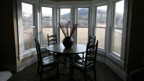 Un comedor con una mesa redonda y cuatro sillas, ventanas amplias con vistas a un paisaje, y un florero con plantas secas sobre la mesa.