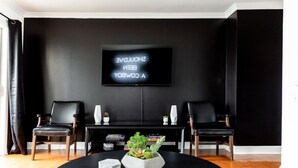 Een moderne woonkamer met een zwarte muur, een flatscreen televisie, twee zwarte leren fauteuils, een salontafel met een plant en een boekenkast met boeken en decoratieve objecten.