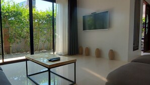 A modern living room with a glass coffee table, a sofa, a flat-screen TV mounted on the wall, and a view of greenery outside. - Ton Yaang Private Pool Villa (Krabi)