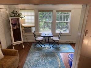 A small room with a table and two chairs, a window with a view of greenery, a radiator, and a cabinet with a mirror.