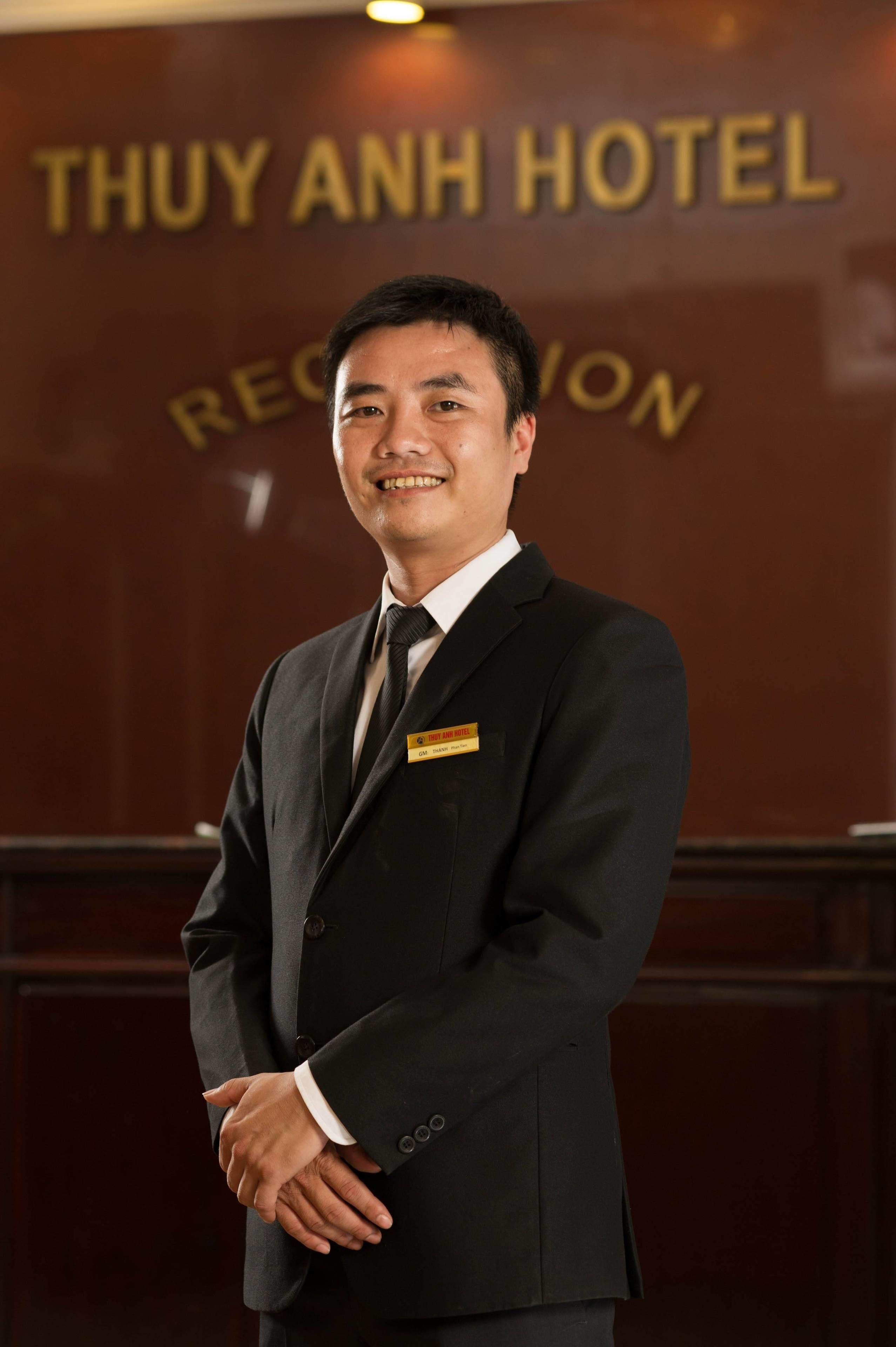Um homem de terno preto com crachá, em frente a uma parede com a inscrição "THUY ANH HOTEL".