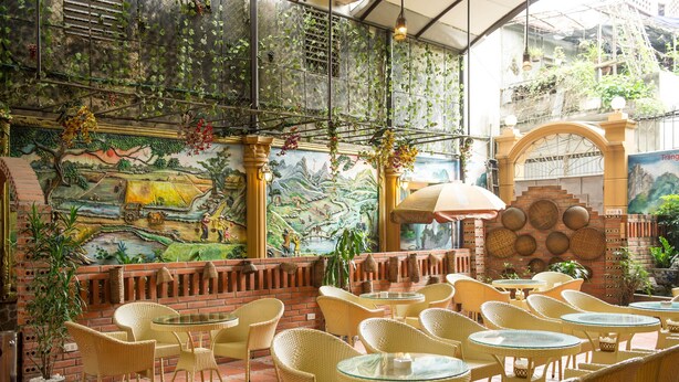 An outdoor cafe with wicker chairs and tables, a mural on the wall, and a brick wall with baskets. - Thuy Anh Hotel (Ninh Binh)