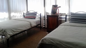 A room with two single beds, a wooden nightstand with a lamp, and a window with sheer curtains. - Soutchai Pakse Backpacker Hostel (Pakse)