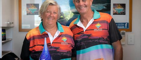 Two people wearing matching shirts with a logo and text, standing in front of a framed picture and a sign with the same logo and text.