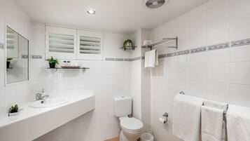 A bathroom with a white sink, toilet, and shower area, with white tiles and a small plant on the counter.