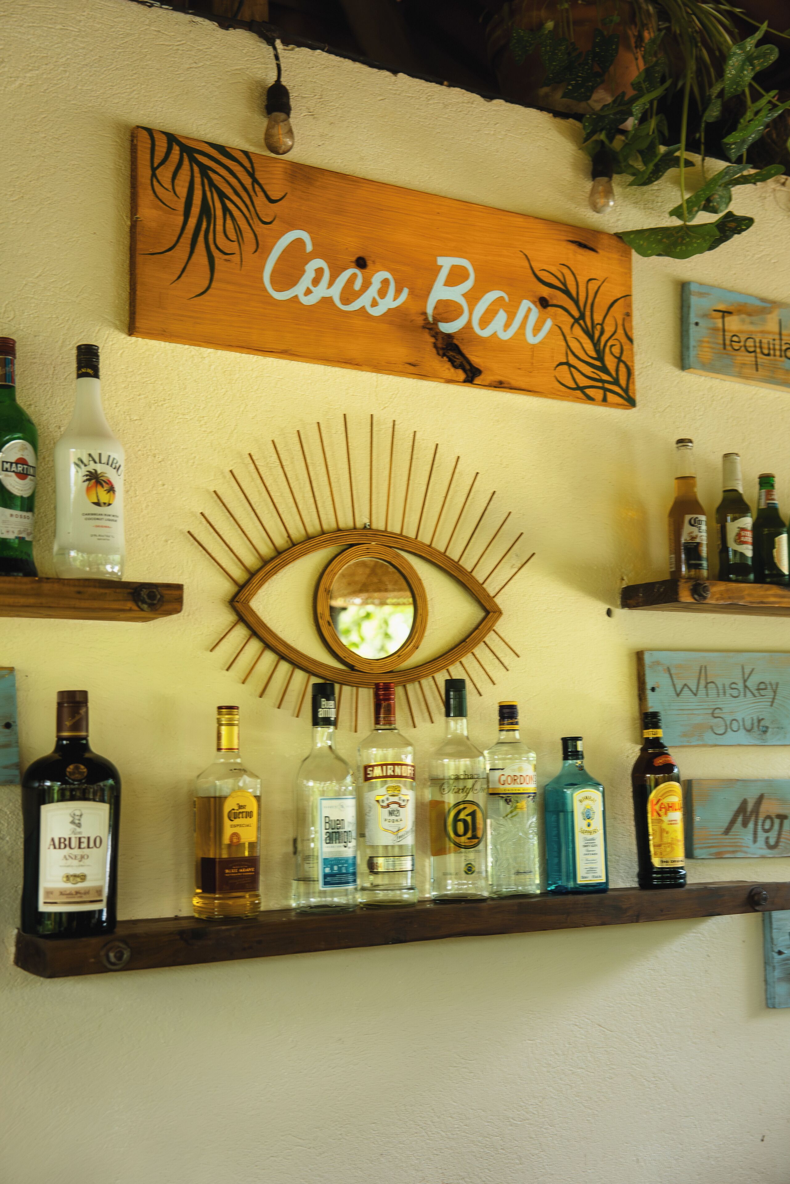 A bar area with a wooden sign reading "Coco Bar," a mirror with an eye design, and shelves with various liquor bottles.