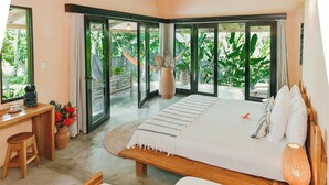 A bedroom with a large bed, a desk, and a view of lush greenery through sliding glass doors. - Villa Coco (Hicaco)