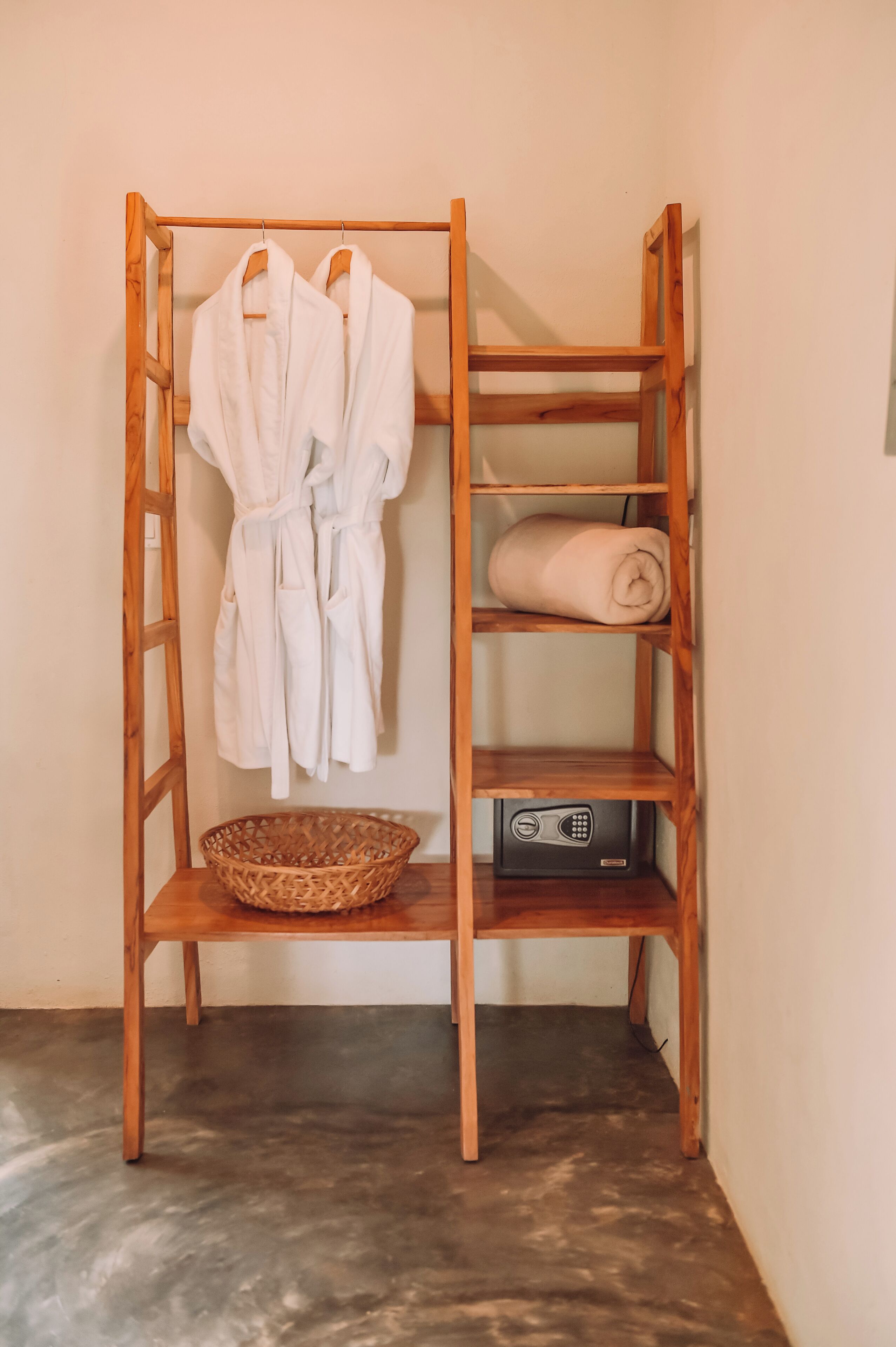 A wooden shelf unit with two white robes hanging on the left and a rolled towel on the right shelf. Below, there is a wicker basket and a black electronic device.