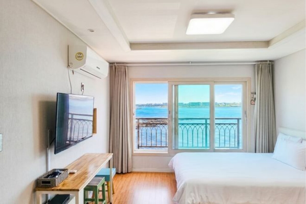 A hotel room with a bed, a TV, a desk, and a window with a view of the sea.
