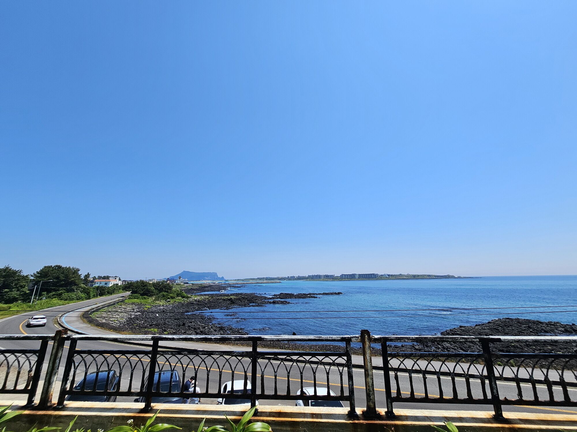 A coastal road with a metal railing, vehicles, and a clear blue sky.