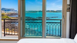 A room with a large sliding glass door leading to a balcony with a view of the ocean and a rocky coastline. - Haemorat (Seogwipo)