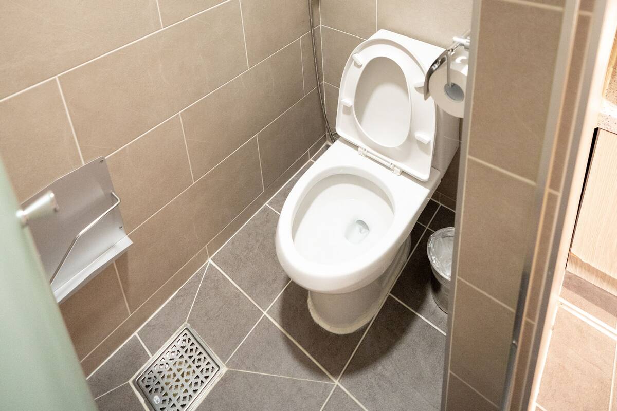 Une petite salle de bain carrelée, équipée de toilettes, d’un sèche-mains et d’une poubelle.