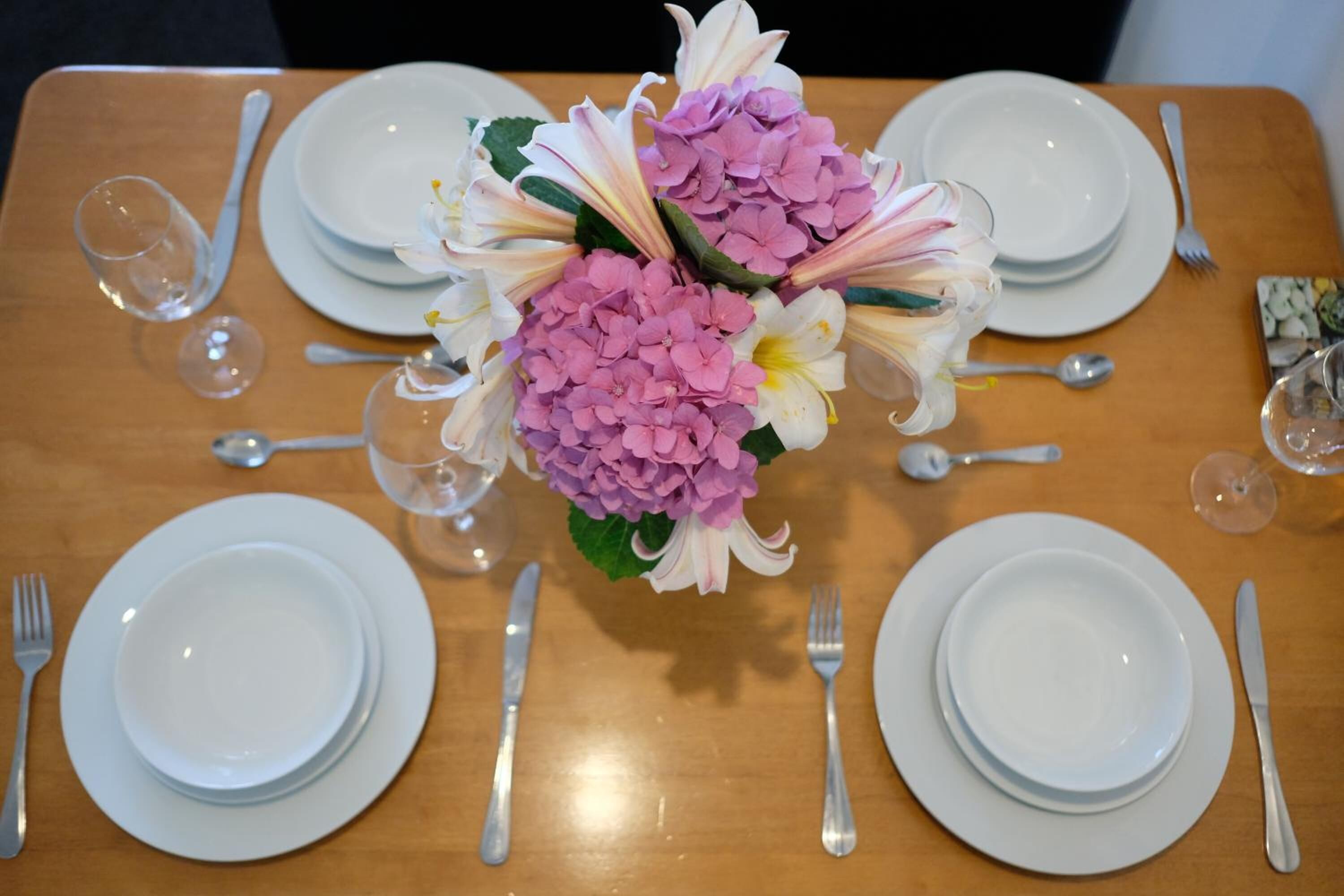 Mesa de comedor dispuesta con platos blancos, cubiertos y un centro de mesa con flores rosas y blancas.