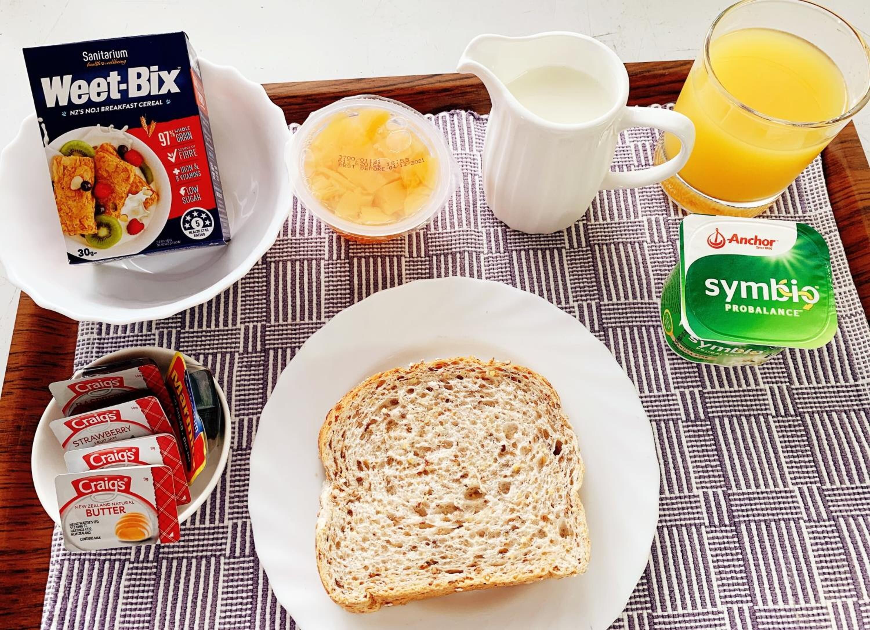 A breakfast tray with Weet-Bix cereal, a glass of orange juice, a jug of milk, a packet of Symbio Probalance yogurt, a slice of bread, and two packets of cream cheese.