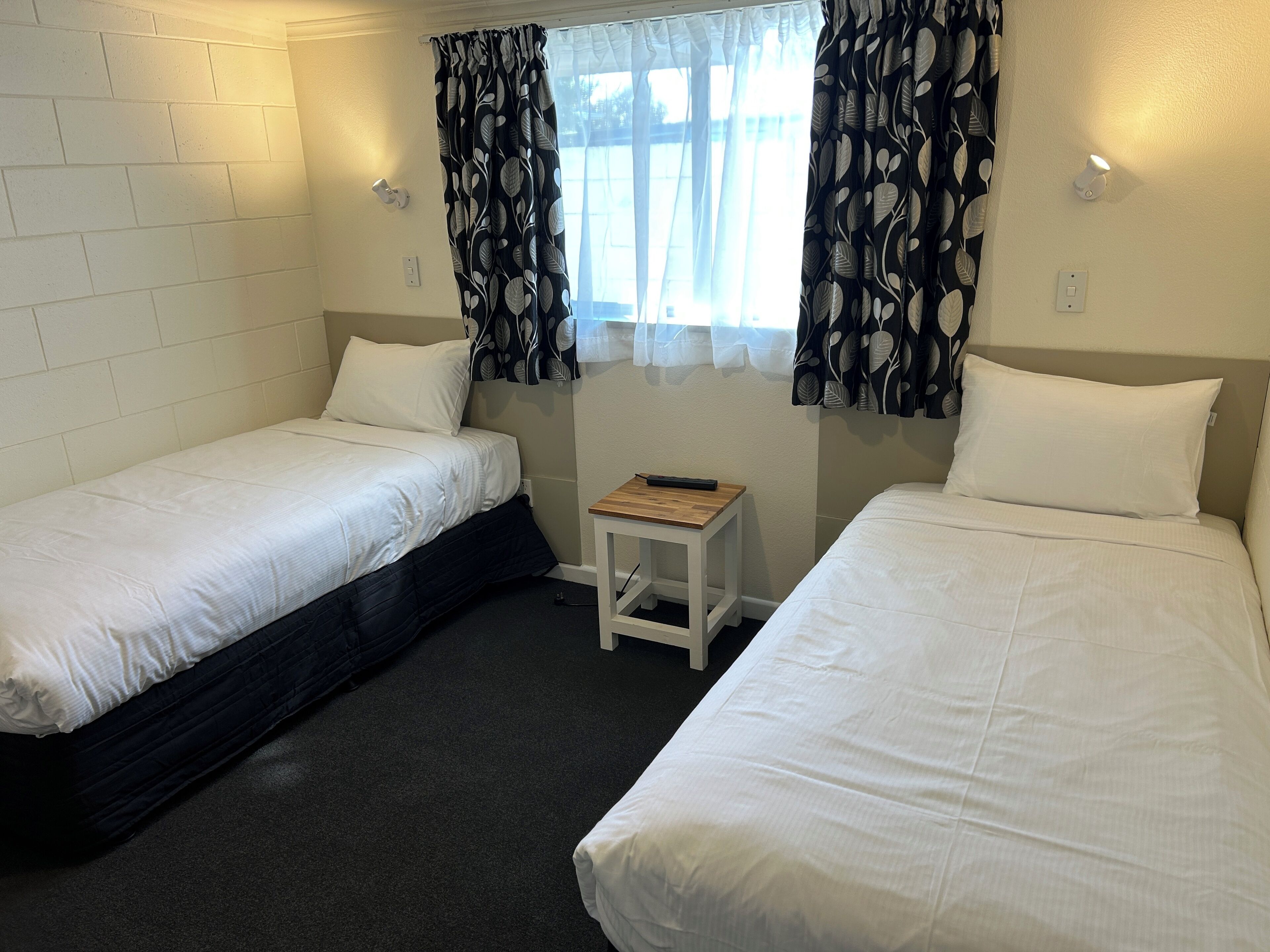 Ein Doppelzimmer mit zwei Betten, einem kleinen Tisch und einem Fenster mit gemusterten Vorhängen.